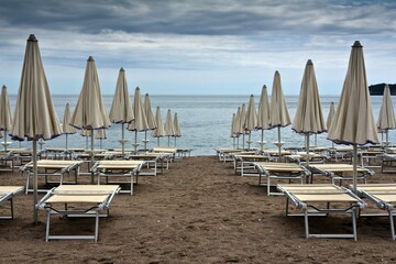 Many identical sunbeds with closed umbrellas on sandy beach of Adriatic sea against cloudy sky in Becici, Montenegro.