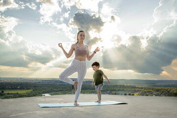 Fototapeta na wymiar Mother and son doing exercise on the balcony in the background of a city during sunrise or sunset, concept of a healthy lifestyle