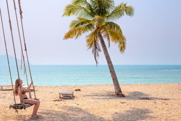 woman in a swimsuit swinging on a swing by the ocean under palm trees, sea vacation and travel.