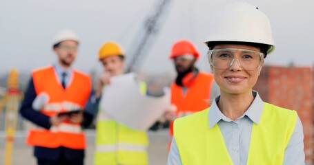 Portrait of beautiful Caucasian young woman in casque and goggles standing at construction site and...