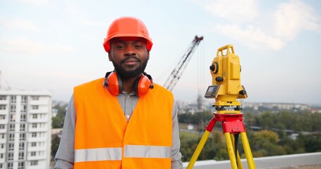 Portrait of handsome young African American man in casque turning to camera at construction site and smiling at total station. Good looking male builder smile at building outside. Topographer.