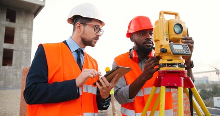 Caucasian man boss in tie with tablet device and in casque. African American male builder measuring angle with total station. Geodesy work. Topography concept. Geodesic job.