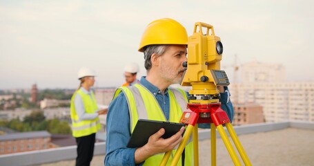 Caucasian man topographer in casque measuring angle with total station on building roof. Male builder constructor doing topographic measures and using tablet device. Geodesic concept. Device computer. - obrazy, fototapety, plakaty