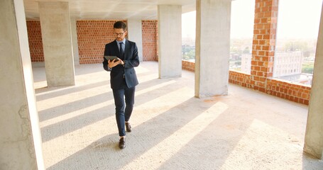 Caucasian male real-estate agent in glasses, suit and tie walking in building on construction phase, using tablet device and tapping or scrolling. Handsome businessman investor choosing mortgage.