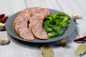 Appetizing sausage sliced on a grey plate on a light grey wooden background with green bell pepper, close up, front view