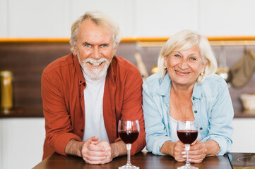 Happy caucasian senior couple stand in the kitchen with wine in their hands and look at the camera smiling