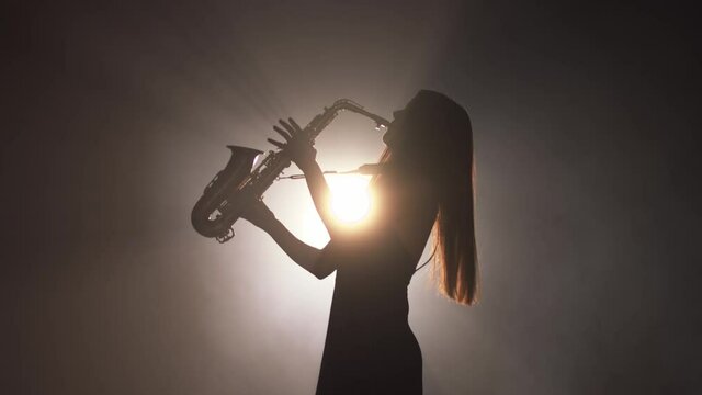 Young beautiful girl in a dark dress plays on a golden shiny saxophone on stage. Dark studio with smoke and stage lighting. Hands and saxophone close up. Side view.Slow motion video