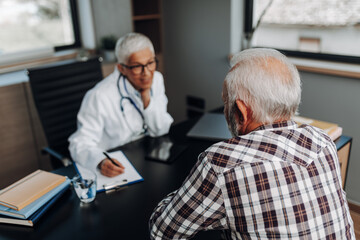 Naklejka na ściany i meble Doctor and senior male patient discussing something while sitting at the table in the doctor's office. Medicine and healthcare concept.