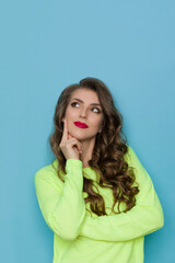 Curious Young Woman In Neon Lime Green Sweater Is Holding Hand On Chin, Looking Up And Thinking.