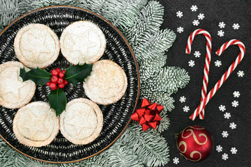 Christmas homemade mince pies on a plate with holly, snow covered fir, candy canes snowflake & bauble decorations on grey grunge background. Festive food composition. Flat lay, top view.