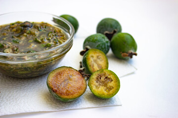 Feijoa jam. Thick, crushed, green in a transparent bowl. A number of large fleshy juicy berries. Ripe dark green with sepals. A scattering of fruit on a light table.