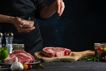 The chef in black uniform pours sea salt on raw pork or beef steak on ingredients background. Frozen motion. Backstage of preparing restaurant delicious food with spices. Concept of cooking process.