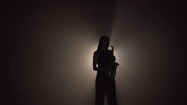 Young beautiful girl in a dark dress plays on a golden shiny saxophone on stage. Dark studio with smoke and stage lighting. Hands and saxophone close up. Side view.Slow motion video