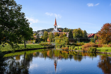 Cityscape view of the village of Schonach in the Black Forest of Germany during autumn. A church is in the centre and reflects in the water of a lake.