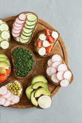Vegetable sandwiches on the wood board on the gray background. Fresh bread with pesto, cucumber, avocado, cherry tomatoes, radish and cheese. Delicious breakfast. Flat lay, top view.