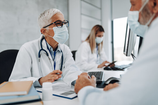 Senior Female Doctor Wears Protective Face Mask While Talking With Patient In Clinic Office During Coronavirus. They Are Sitting At The Table In The Doctor's Office. Pandemic, Covid-19 Concept.