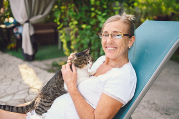 Happy smiling senior woman in glasses relaxing in summer garden outdoors hugging domestic tabby cat