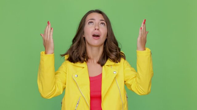 Angry dissatisfied young brunette woman 20s years old in casual yellow leather jacket posing isolated on green background studio. People lifestyle concept. Looking up ask why spreading hands swearing