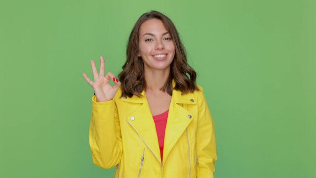 Cheerful funny young brunette woman 20s years old in casual yellow leather jacket posing isolated on green background in studio. People lifestyle concept. Looking camera showing Ok gesture blinking