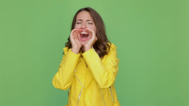 Crazy young brunette woman 20s years old in casual yellow leather jacket posing isolated on green background in studio. People lifestyle concept. Looking aside screaming with hands gesture near mouth