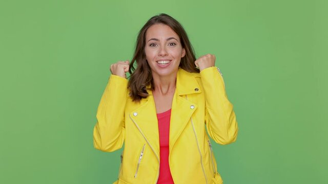 Side profile view of surprised young woman 20s in yellow leather jacket isolated on green background studio. People lifestyle concept. Turn around camera say wow doing winner gesture showing thumbs up