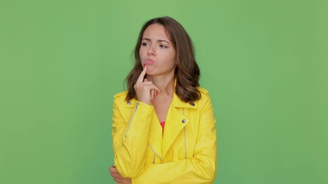 Displeased confused boring young brunette woman in yellow leather jacket posing isolated on green background studio. People lifestyle concept. Looking aside put hand prop up on chin spreading arms