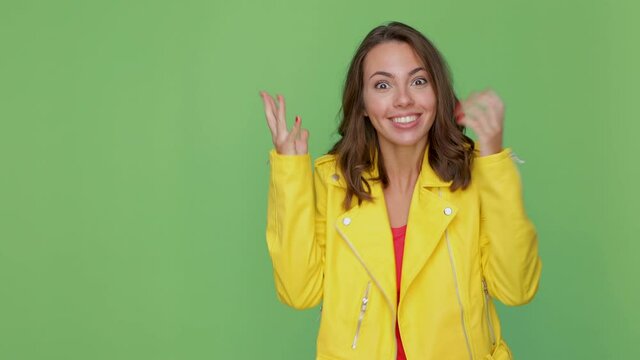 Funny excited young brunette woman 20s in yellow leather jacket posing isolated on green background studio. People lifestyle concept. Point index finger aside say wow spreading hands showing thumbs up