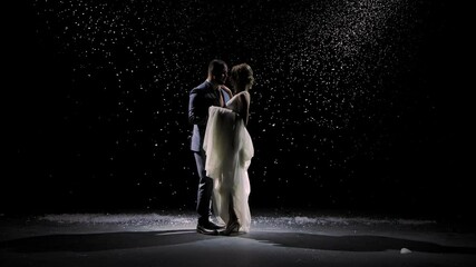 A charming couple in love dances against the background of falling snow. A man in a suit and a woman in a white long dress on a black background in the rays of studio light. Slow motion.