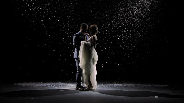 A charming couple in love dances against the background of falling snow. A man in a suit and a woman in a white long dress on a black background in the rays of studio light. Slow motion.
