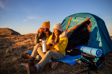 Two happy girls drinking hot tea at the front of the tent and camping in the mountains.
