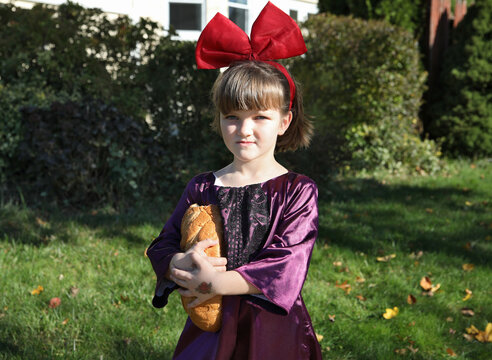 Girl in Halloween costume in violet dress and big red bow, with a loaf of white bread looking at camera.
