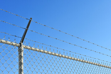 An abstract image of a tall industrial fence with barbed wire against a bright blue sky. 