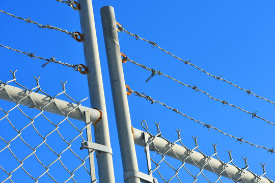 An abstract image of a tall industrial fence with barbed wire against a bright blue sky. 