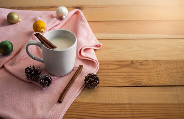 Hot drink for autumn winter concept. Hot milk with cinnamon in white cup and pine cones, Christmas decorations, sweater on wooden background.