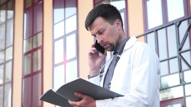 Healthcare professional makes phone call while reading patient chart near hospital. Young male doctor holding a folder with papers and talking on the phone. Telemedicine, telehealth concept