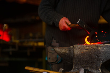 A close-up of the hands of a forge with a hammer striking a red-hot metal workpiece lying on an anvil and burning sparks scatter around