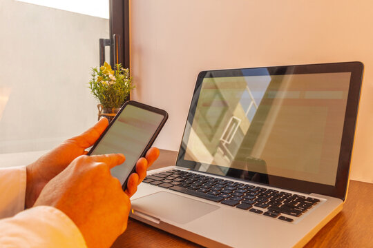 hands of a man navigating through his cell phone with a laptop computer coordinated with the same website. Image in warm tones