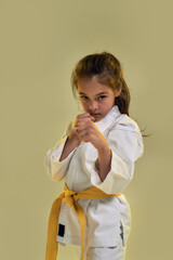 Portrait of little karate girl in white kimono with a yellow sash looking focused at camera, fighting, doing martial arts while standing isolated over yellow background © Svitlana