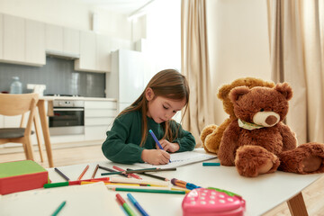 A cute little girl writing with a marker having her teddybear around sitting at a table in a huge bright kitchen