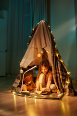 A little girl wearing pyjamas sitting on a floor barefoot in a self-made hut made of a plaid...