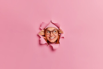 Smiling glad freckled young woman wears optical glasses and looks away notices something interesting has white teeth breaks head through torn paper pink studio background. Happy emotions concept