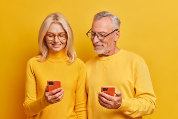 Middle aged application devoted couple check information on their cellulars watch video and photos on modern gadgets communicate with grown up children isolated over yellow background. Chatting online