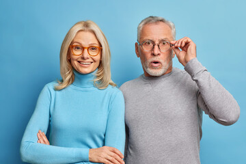 Fototapeta Cheerful middle aged woman and her husband pensioner reacts on shocking news keeps hand on spectacles isolated over blue background. Family age and emotions concept. Senior couple at pose home obraz