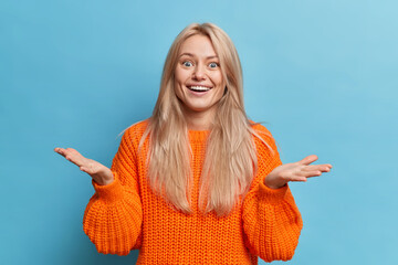 Cheerful unaware European woman spreads palms with bewilderment cannot believe in success stands indoor wears orange knitted sweater isolated over blue studio background. Human reactions concept © WHstudio Leushin N