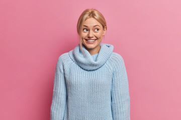 Satisfied blonde woman has dreamy satisfied expression dressed in blue knitted jumper looks away gladfully isolated over pink background. Happy glad European lady considers to buy something.