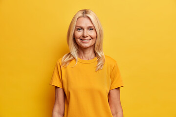 Pretty blonde middle aged woman smiles gently dressed in casual clothing expresses positive emotions. Cheerful European female enjoys pleasant talk isolated over yellow background. People age concept