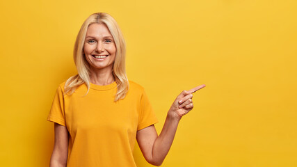 Portrait of pleasant looking blonde woman with satisfied expression points at blank space dressed...