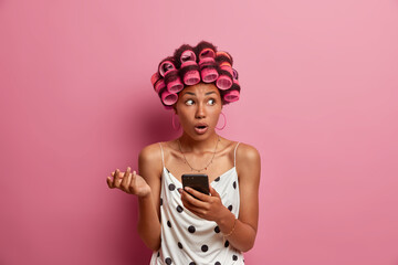 Beautiful embarrassed surprised dark skinned Afro American woman holds mobile phone and looks with puzzlement aside dressed in polka dotted dress applies hair rollers for making perfect hairstyle