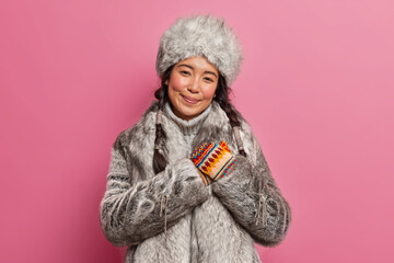 Pleased brunette woman inhabitant of far north makes gratitude gesture smiles gently wears grey fur winter outfit poses against rosy background. Touched arctic lady from far north being thankful