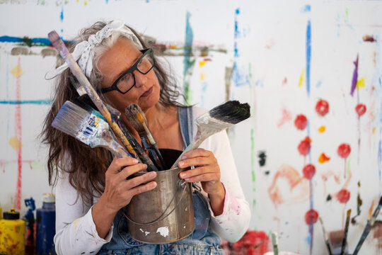 Older Woman, Artist, In Her Fifties With Grey Hair And Black Glasses And Many Paintbrushes, Copy Space